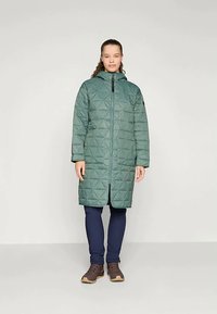 Moteris, apsivilkusi ilgą žalią siūtuotą striukę su kapišonu, tamsiai mėlynus kelnes ir rudas žygių batus, stovi prieš paprastą šviesų foną.