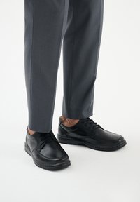 Scarpe in pelle nera con una superficie liscia, design con lacci e punta arrotondata. Abbinate a pantaloni grigi su misura.