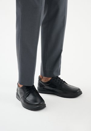 Schwarze Lederschuhe mit glatter Textur, Schnürdesign und abgerundeter Zehe. Kombiniert mit maßgeschneiderten grauen Hosen.