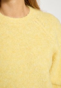 Dzeltena adīta džemperis ar pūkainu tekstūru, apaļu kakla izgriezumu un raglāna piedurknēm. Audums izskatās mīksts un viegls, ar smalku rakstu.