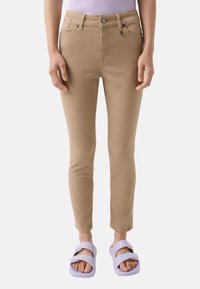 Beige fitted jeans with five pockets, a zip fly, and a button closure. Paired with light purple slide sandals. Smooth texture.