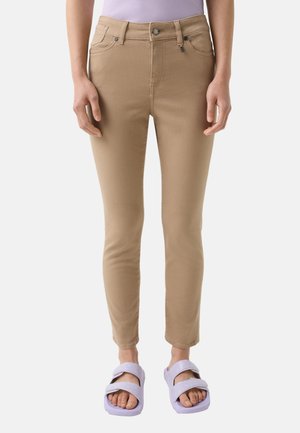 Beige enge Jeans mit fünf Taschen, einem Reißverschluss und einem Knopfverschluss. Kombiniert mit helllila Rutschen-Sandalen. Glatte Textur.