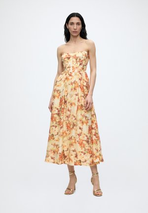 Vrouw met lang donker haar die een strapless, bloemen, midi-lengte jurk draagt in warme herfstkleuren en gouden sandalen met hoge hakken en bandjes.