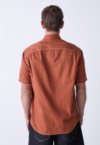 Short-sleeve shirt in rust-orange fabric with a textured finish. Features a classic collar and stitched detailing along the shoulders.