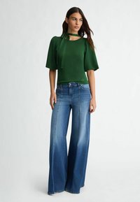 Top verde lavorato a maglia con maniche a sbuffo corte e dettaglio a fiocco, abbinato a jeans in denim a gamba larga di colore blu chiaro. Tessuto morbido e stile casual.