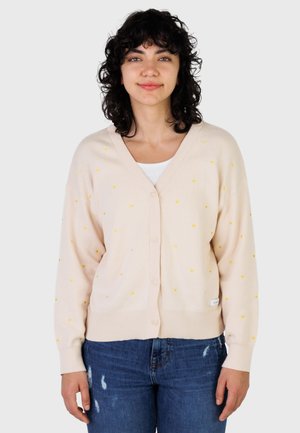 Kvinne med krøllete mørkt hår som har på seg en beige cardigan med små gule hjerte mønstre over en hvit topp og blå slitte jeans.