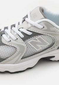 Chaussure de course grise et blanche avec un dessus en mesh, une languette rembourrée et une semelle en caoutchouc contrastée avec des accents texturés. Logo "N" visible.