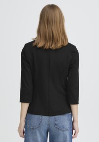 Nainen, jolla on olkapäihin ulottuvat vaaleanruskeat hiukset, yllään musta bleiseri ja siniset farkut, esitettynä takaa yksinkertaisen taustan edessä.