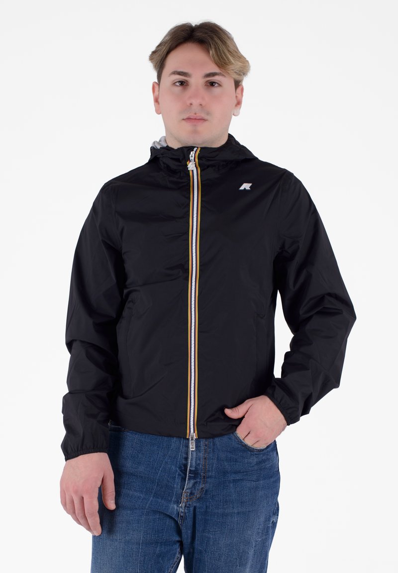 Giacca leggera nera con cappuccio, con cerniera bianca e gialla e un piccolo logo sul petto. Indossata con jeans blu.