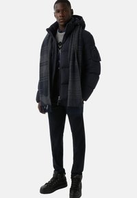 Granatowa pikowana kurtka z kapturem, szary szalik w kratę, ciemne spodnie i czarne buty na sznurowadła. Gładka faktura i elegancki design.