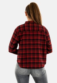 Donna con capelli lunghi raccolti in un alto chignon, che indossa una camicia a quadri rossi e neri e jeans grigi sbiaditi, vista da dietro su uno sfondo bianco.