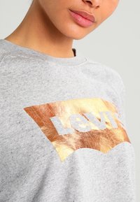 Sweatshirt gris avec un graphique en cuivre métallique en forme de bouclier portant le mot « Levi's » en tissu texturé blanc.