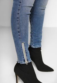 Tunna blå denimjeans med siddragkedjor, kombinerade med spetsiga svarta högklackade skor som har en slät textur och en formsydd design.