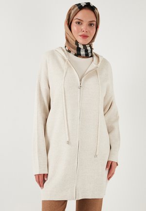 Lichtbeige rits-hoodie met trekkoorden, zachte textuur, lange mouwen en capuchon. Draagt over een crème shirt, gecombineerd met bruine broek.