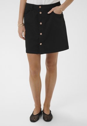 Femme portant une jupe noire boutonnée sur le devant avec des poches, associée à des ballerines tissées noires et des bracelets dorés aux poignets.