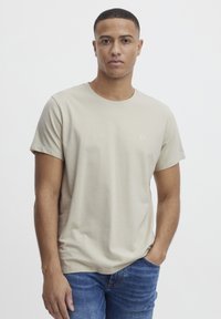 Beige bomull t-skjorte med rund hals og korte ermer. Har en liten logo på brystet. Brukt med blå jeans.