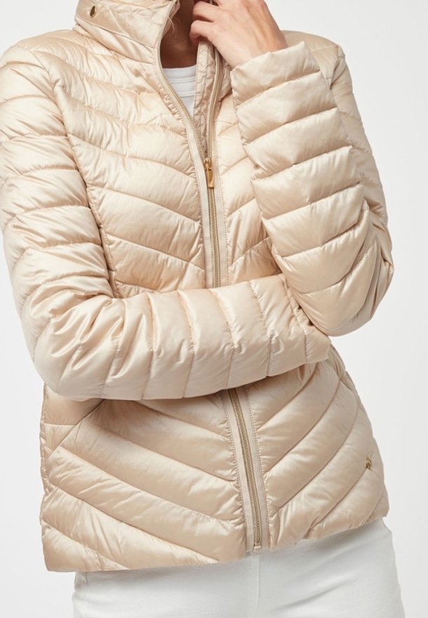 Doudoune matelassée beige avec fermeture éclair devant, portée par une personne les bras croisés et touchant son visage, vêtue d’un pantalon blanc.