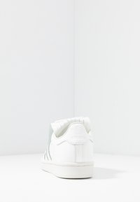 Vit sneaker med en texturerad baksida, utrustad med en ljusgrå panel och tre mörkgrå ränder. Gummisula och präglad logotyp på hälen.