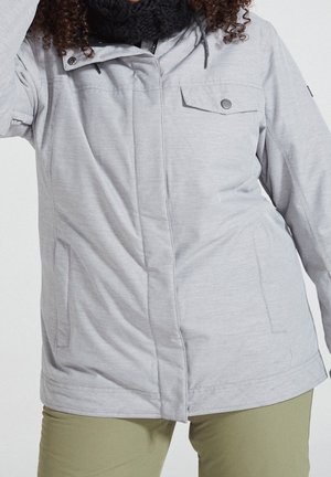 Gros plan sur une personne portant une veste zippée gris clair avec poche à rabat et un pantalon vert olive, assortis à une écharpe en tricot noire.