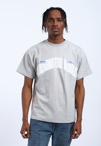 Camiseta de algodón gris de manga corta con grandes letras blancas "AROUND THE WORLD" y acentos azules. El modelo lleva puestos unos vaqueros de mezclilla azul.
