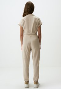 Jumpsuit beige con maniche corte, vita elastica e caviglie con risvolto. Tessuto morbido e leggero; design minimalista senza motivi visibili.