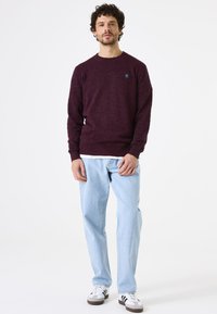Bordeaux pullovertrui met een ronde hals, een gestructureerde breiwerk en een klein logo op de borst, gecombineerd met lichtblauwe jeans en sneakers.