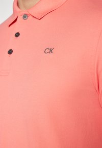 Ružová polo tričko vyrobené z textúrovanej látky s trojitou gombíkovou légou a vyšitým logom "CK" v kontrastnej farbe.