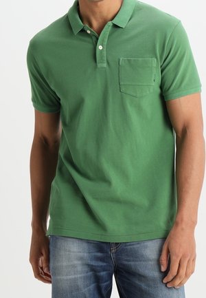 Mann trägt ein grünes kurzärmeliges Polohemd mit Brusttasche und blaue Jeans, steht vor einem weißen Hintergrund.