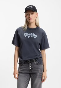 Tmavě šedé tričko s grafickým motivem ve tvaru srdce, na kterém je napsáno "HUGO" světle šedou barvou, doplněné o šedé džíny a černou čepici s názvem značky.