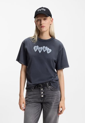 Dark gray T-shirt with heart-shaped graphic spelling "HUGO" in light gray, paired with gray jeans and a black cap featuring the brand name.