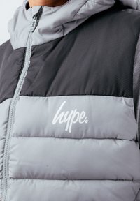 Grå vadderad jacka med svart överdel, hexagonalt mönster, dragkedja framtill och vitt logo på vänstra bröstområdet.