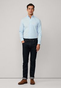 Camicia azzurro chiaro con bottoni, jeans di denim scuro, scarpe in suede marrone; design aderente con maniche lunghe e polsini arrotolati. Stile minimalista.