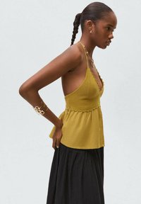 Top sans manches jaune moutarde avec un décolleté en forme de vagues, un buste texturé et un peplum froncé, associé à une jupe noire fluide.