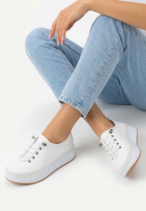 Person wearing light blue jeans and white lace-up platform shoes, sitting with legs crossed on a white surface.