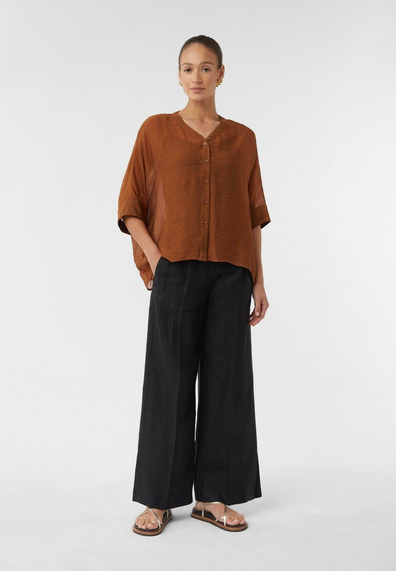 Frau steht auf einem weißen Hintergrund und trägt eine lockere, rostfarbene Bluse mit Knöpfen, weite schwarze Hose und goldene, geschnallte flache Sandalen.