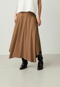 Hnědá midi sukně s jemným designem vyrobená z měkké látky. V kombinaci s černými klínovými botami, které mají lesklou texturu a špičatou špičku.