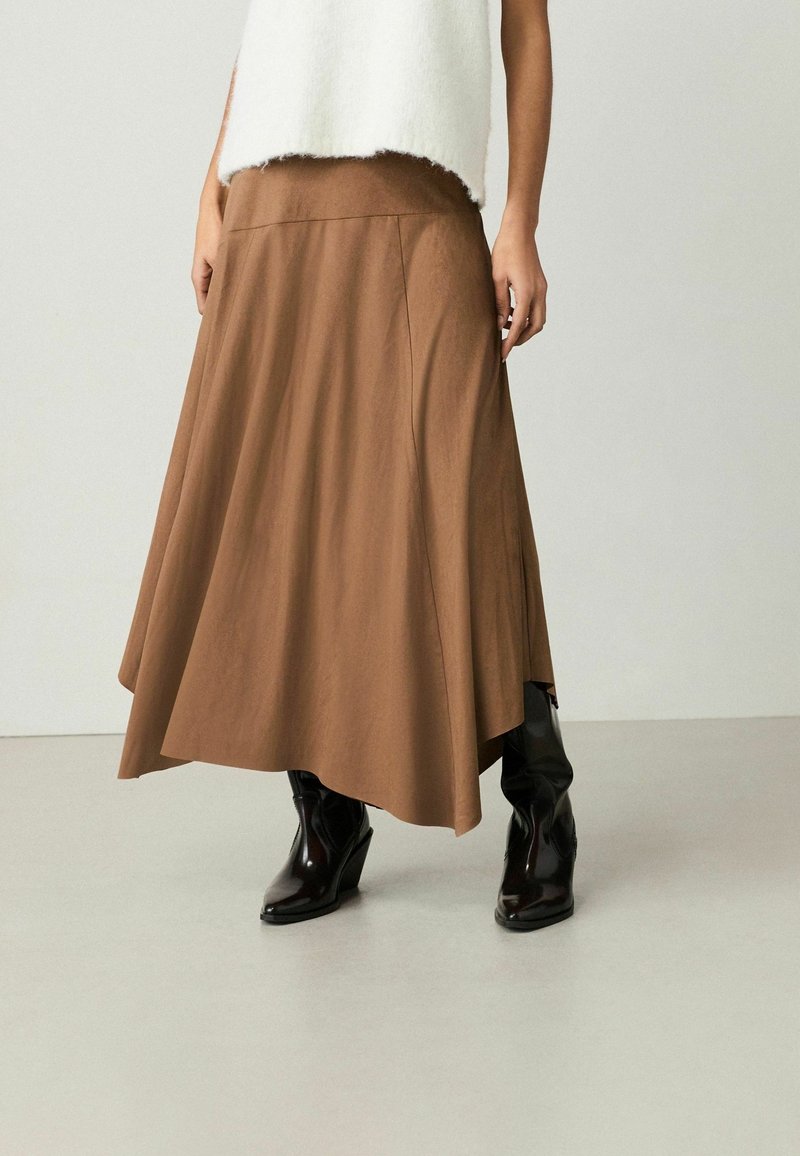 Hnědá midi sukně s jemným designem vyrobená z měkké látky. V kombinaci s černými klínovými botami, které mají lesklou texturu a špičatou špičku.