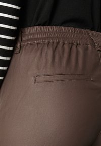 Pantalons extensibles marron avec une taille élastique, dotés d'une poche arrière unique et d'une texture lisse, associés à un haut rayé noir et blanc.