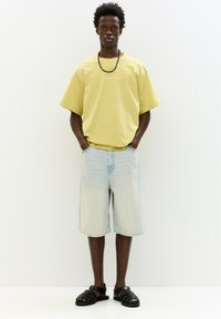 Žluté oversized tričko s krátkými rukávy, světle modré džíny šortky a černé sandály na pásky. Jednoduchý, neformální outfit bez vzorů.