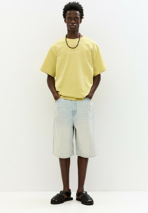 Geel oversized T-shirt met korte mouwen, een paar lichtblauwe denimshorts en zwarte sandalen met banden. Eenvoudige, casual outfit zonder patronen.