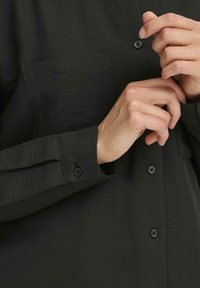 Hands buttoning a black long-sleeve shirt with visible buttons and a front pocket, close-up on torso and arms.