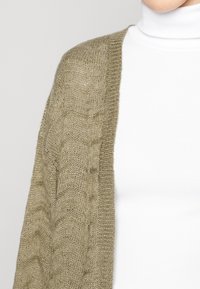 Olivgrüner Strickcardigan mit strukturiertem Muster und offener Vorderseite, über einem weißen Rollkragenoberteil getragen. Weiches, leichtes Material.