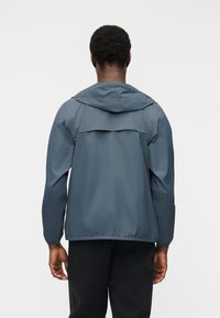 Άνδρας με μπλε ζακέτα με κουκούλα, φαίνεται από πίσω, στέκεται μπροστά σε απλό λευκό φόντο.