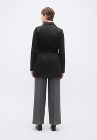 Μαύρο μπουφάν με ζώνη και κομψό σχεδιασμό, με μακριά μανίκια και λεία υφή, συνδυασμένο με γκρι παντελόνι φαρδιάς γραμμής.