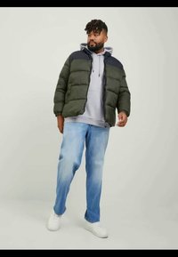 Doudoune verte olive avec un col bleu marine, associée à un sweat à capuche gris et un jean en denim bleu clair. Modèle portant des baskets blanches.