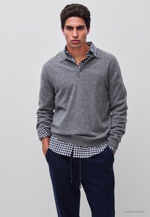 Homme portant un pull gris par-dessus une chemise à carreaux noirs et blancs avec un pantalon bleu marine, debout les mains dans les poches devant un fond uni.