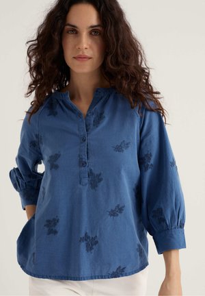 Femeie cu păr creț și închis la culoare, purtând o bluză albastră cu mâneci lungi și cu modele florale brodate, precum și pantaloni albi, pe un fundal simplu.