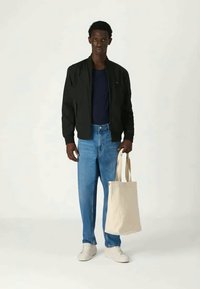 Μαύρο bomber jacket, μπλε μπλουζάκι, ανοιχτόχρωμο τζιν, λευκά sneakers και μπεζ τσάντα tote, στέκεται μπροστά από ένα ουδέτερο φόντο.