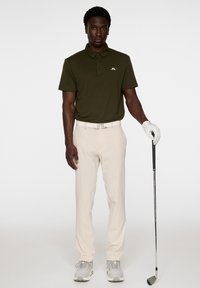 Golfowa koszulka polo w ciemnym oliwkowym kolorze z logiem z przodu, w połączeniu z jasnobeżowymi spodniami, białym paskiem, rękawiczkami i szarymi butami, trzymająca kij golfowy.