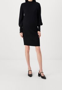 Robe noire en maille côtelée avec de longues manches bouffantes et un col haut, associée à des escarpins noirs à bout pointu et à bride arrière.